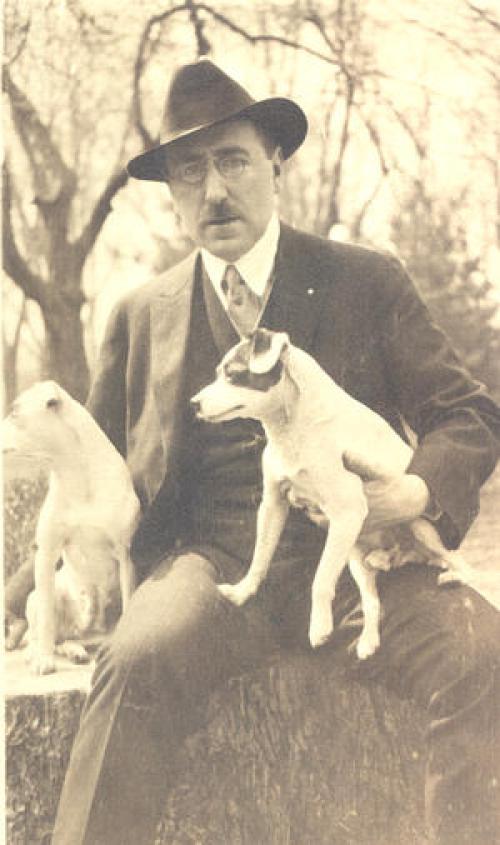A black and white portrait of Twitmeyer in a suit holding two dogs and sitting outside near a forest